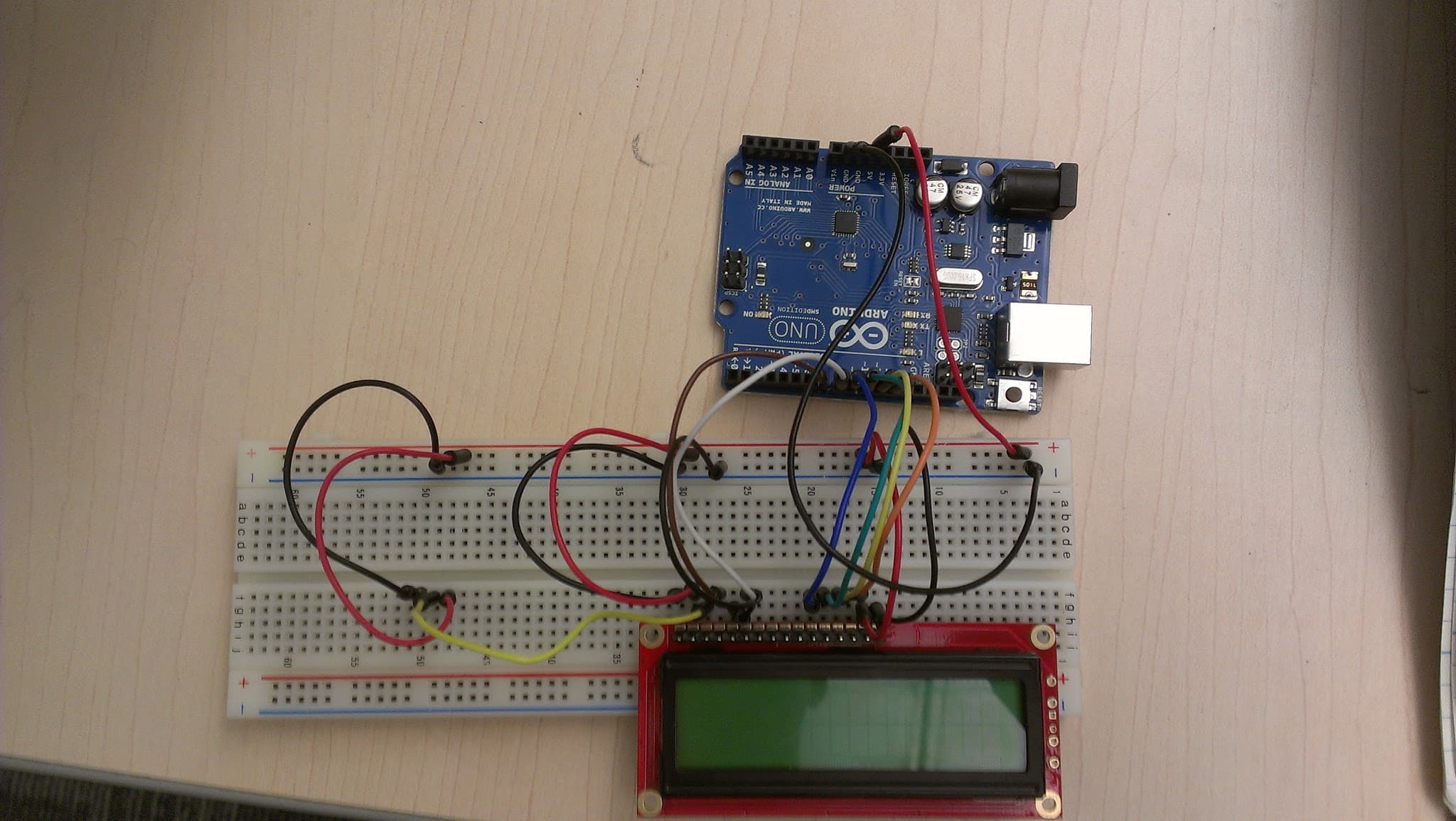 An Arduino Uno board on a wooden desk connected by coloured jumper leads across a white solderless breadboard to a red 16 by 2 character LCD module with a green backlit display, with no casing and no soldered joints