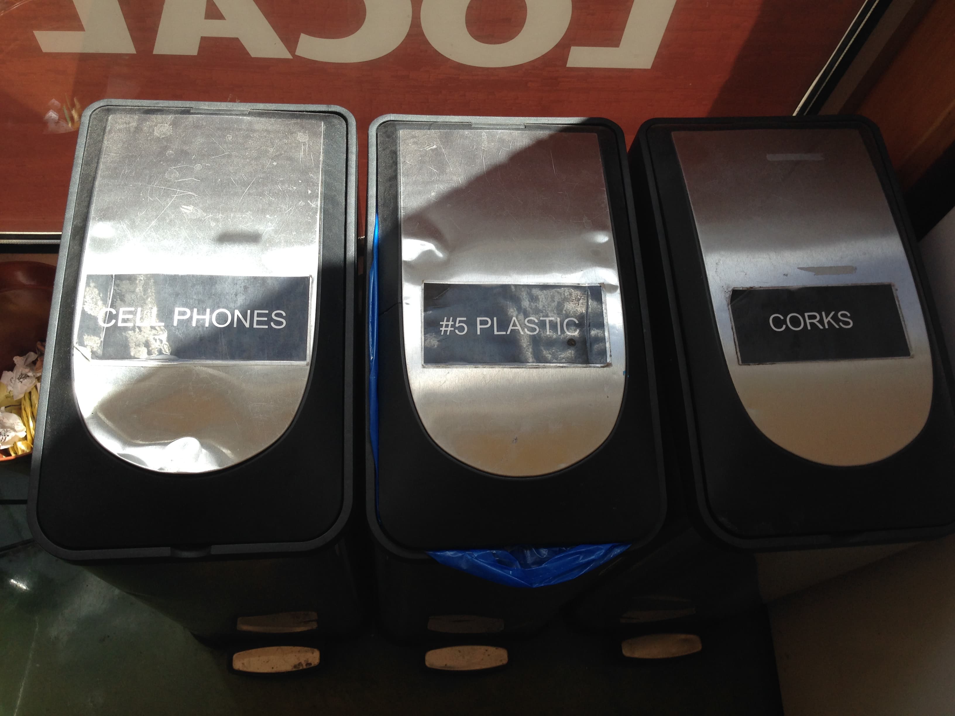 A row of metal-topped recycling bins against a red wall, the nearest clearly labelled CELL PHONES, the others labelled for plastic and corks