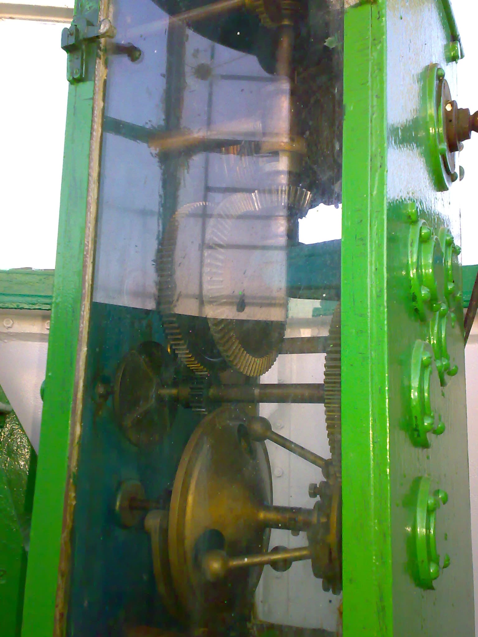 A mechanism of meshing brass and steel gears of different sizes behind a glass guard in a green painted frame, part of a lighthouse lens rotation drive