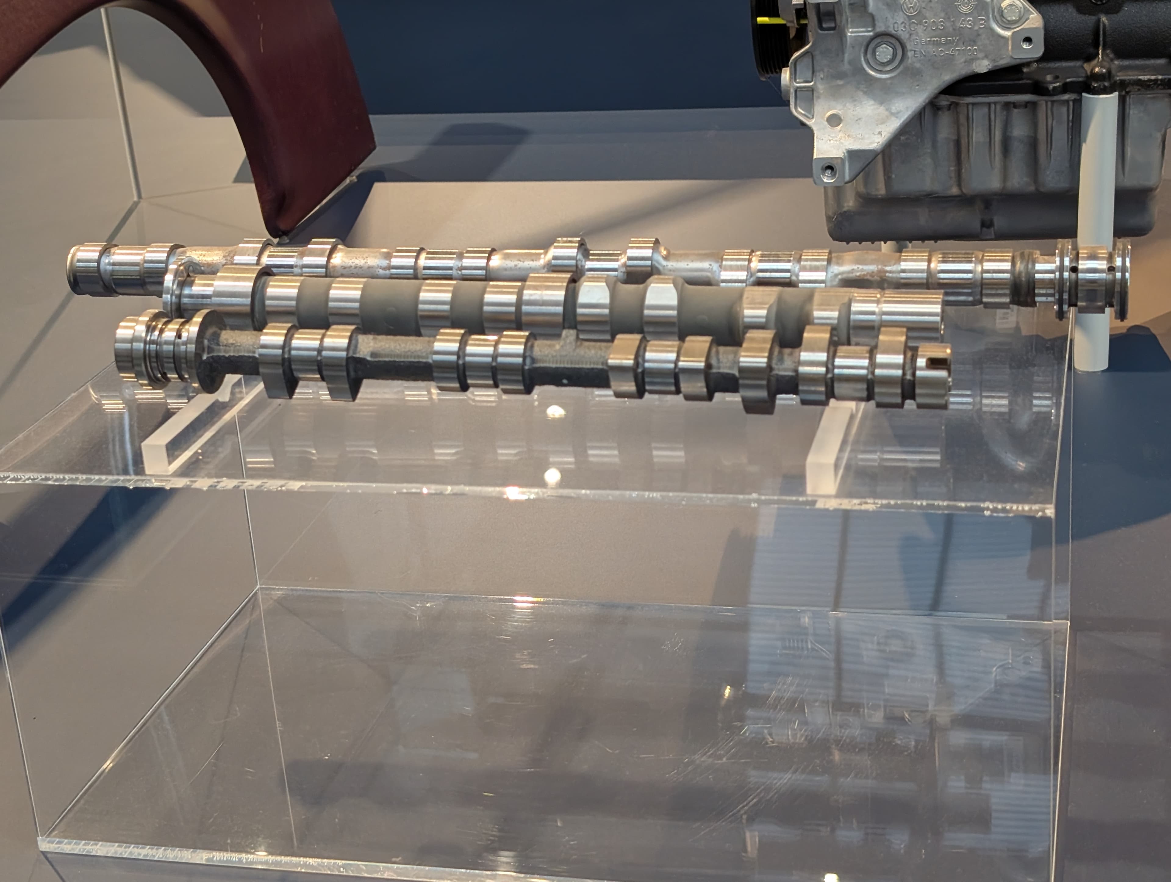 Three steel camshafts displayed side by side in a glass case, each a long shaft with a series of shaped lobes and circular bearing journals spaced along its length