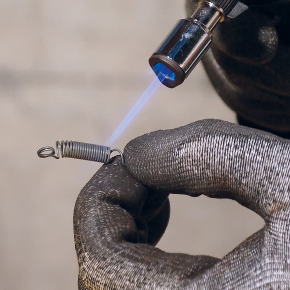 A gloved hand holding a small metal spring while a blue torch flame is played over it, the spring recovering its original coiled shape as it heats